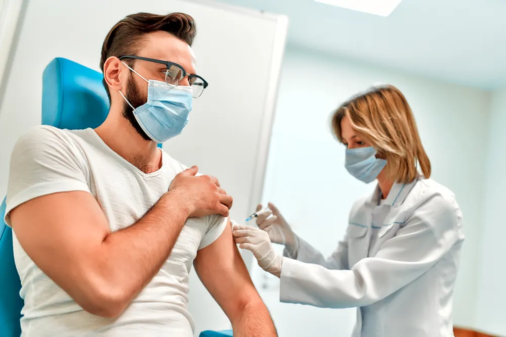 Vital Plus Pflegedienst - Junger Mann mit medizinischer Gesichtsmaske lässt sich in einer Arztpraxis gegen Covid-19 impfen