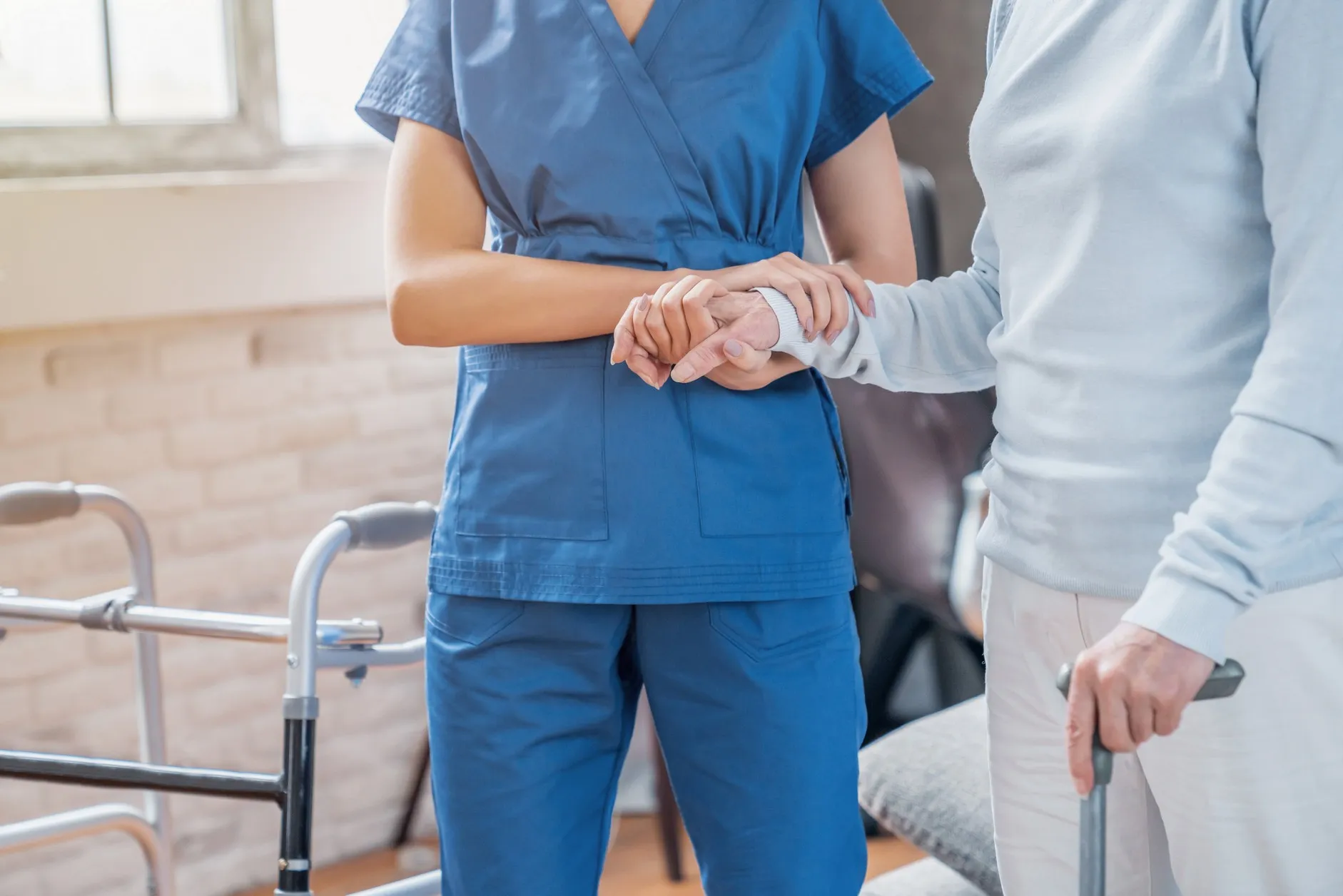 Vital Plus Pflegedienst - Ausschnitt einer Sozialarbeiterin, die einer älteren Frau zu Hause beim Gehen mit dem Rollator hilft
