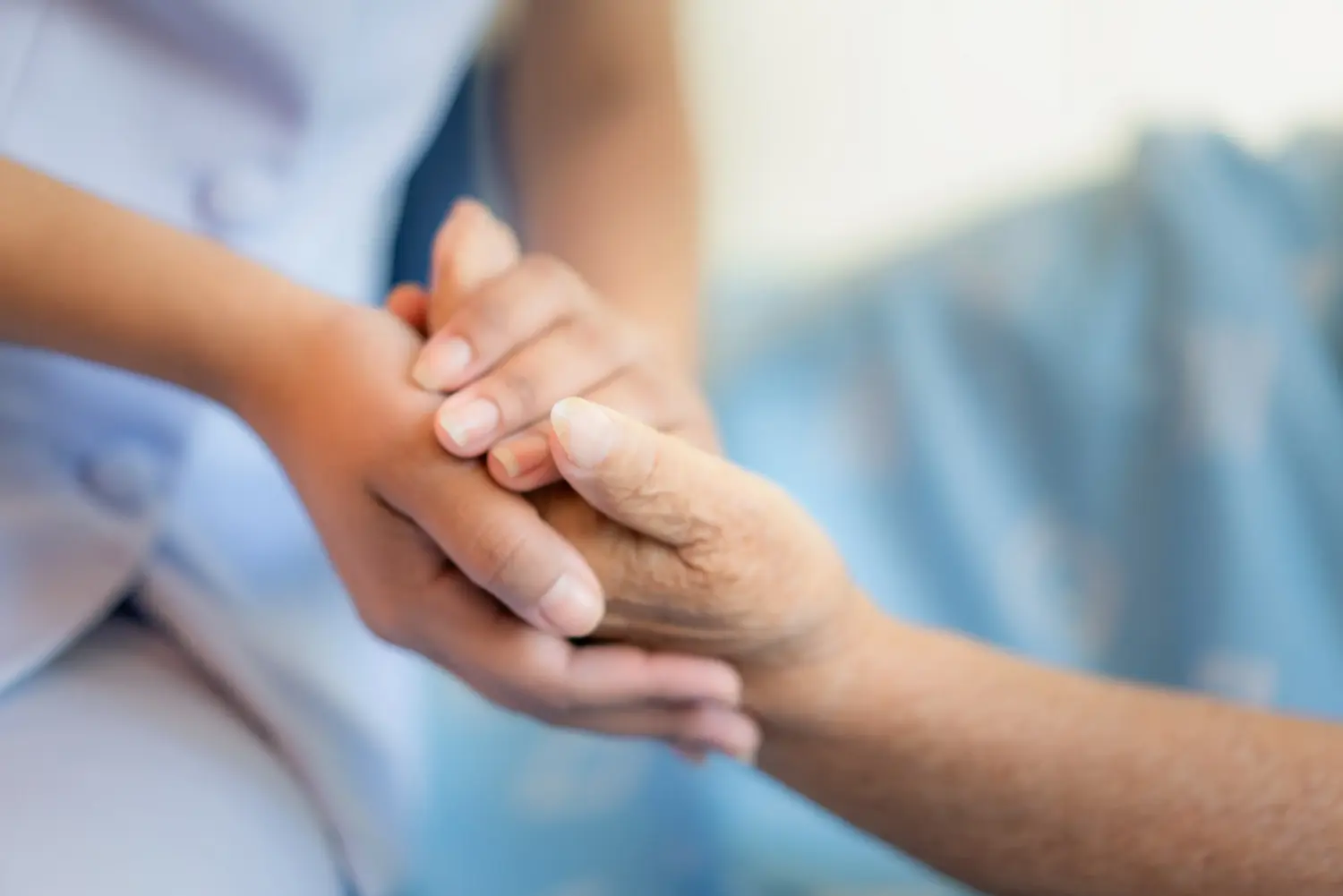 Vital Plus Pflegedienst - Krankenschwester auf einem Krankenhausbett sitzend neben einer älteren Frau mit helfenden Händen, Konzept für die Altenpflege